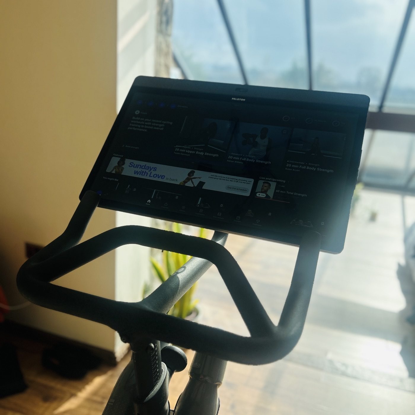 A Peloton bike's touchscreen showing a personalised recommendation to build on recent cycling workouts with strength training, alongside thumbnails of 20-minute strength classes.